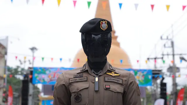 Robô policial com uniforme militar e rosto coberto por visor preto, em evento público na Tailândia, com bandeirolas coloridas ao fundo.