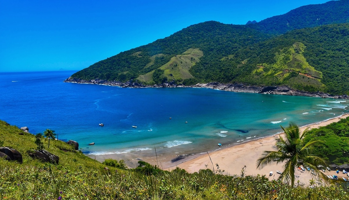 Vista panorâmica de uma praia tropical com mar azul, montanhas verdes e barcos ancorados