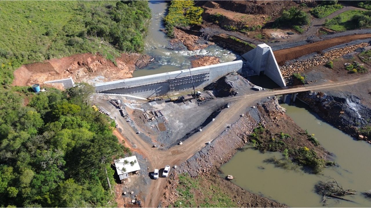 Imagem aérea de uma hidrelétrica em construção em Santa Catarina, mostrando a barragem, máquinas de obra, áreas escavadas e o rio passando pela estrutura.