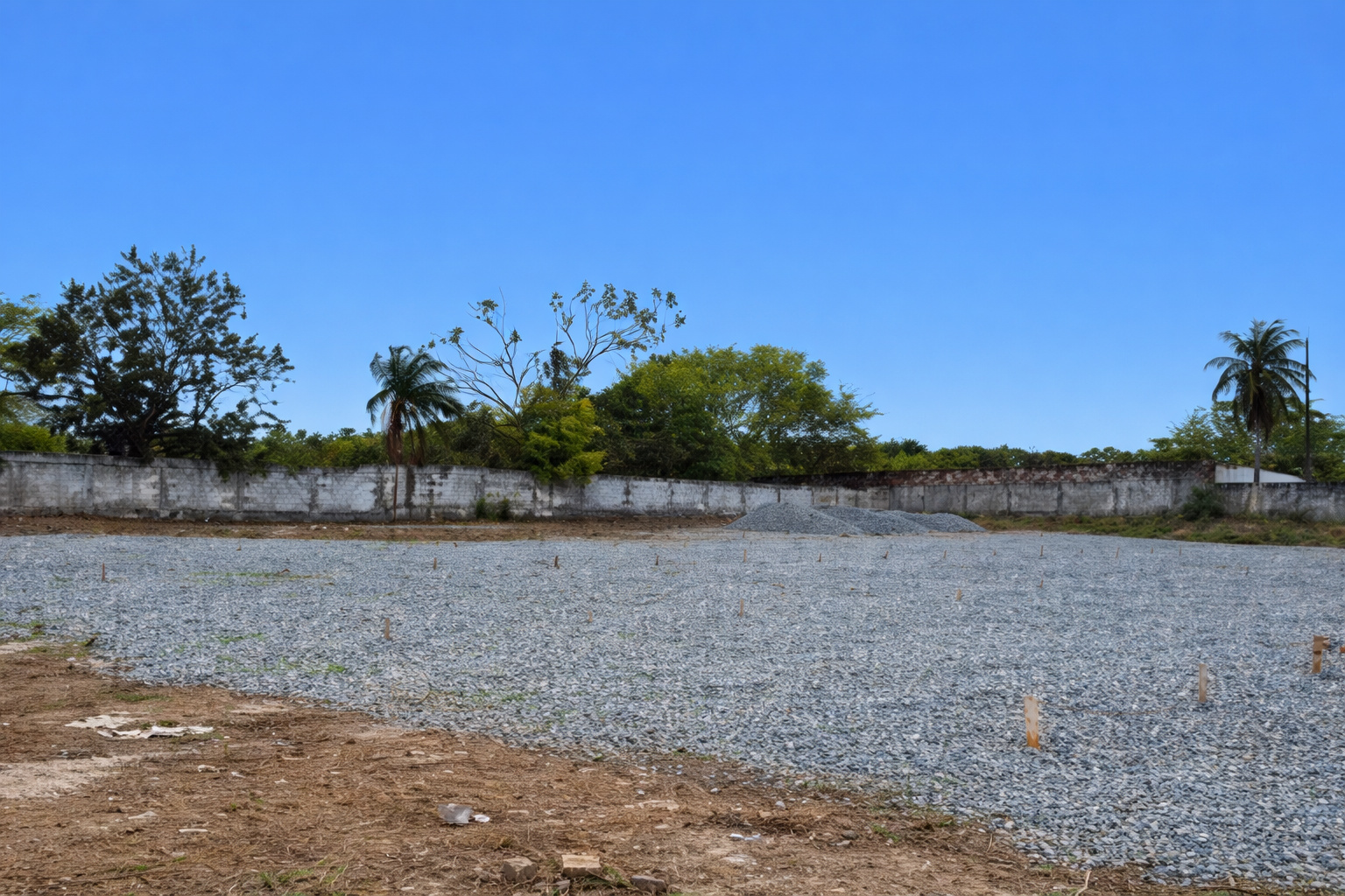 Terreno preparado para construção, coberto com brita, cercado por muro e árvores, sob céu azul limpo.