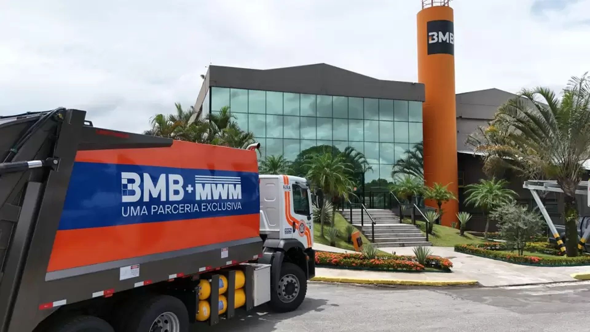 Caminhão de coleta de resíduos com identidade BMB + MWM movido a gás estacionado em frente a prédio corporativo moderno com fachada de vidro e torre laranja.